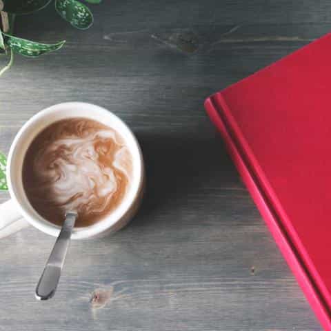 A silver spoon in a white cup of coffee sitting next to a red book and a plant on a desk