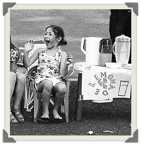 Anamaría Morales as a child wears a white dress and smiles, sitting beside a lemonade stand with pitchers of lemonade, Photo Credit: Anamaría Morales
