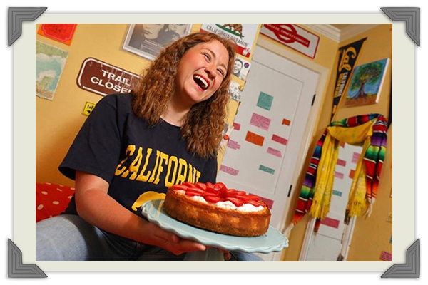 Anamaría Morales, una joven con el cabello rizado y castaño, vistiendo una camiseta negra con la palabra California y sonriendo con un pay de queso en las manos. Crédito fotográfico: Anamaría Morales
