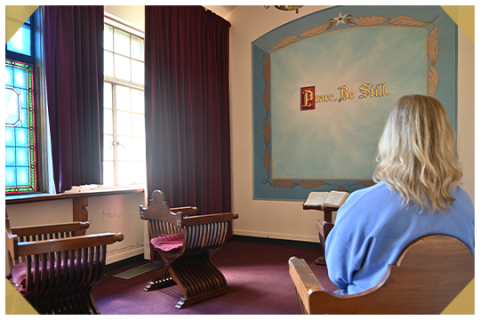 A blonde woman sitting in the Peace Chapel with a large painting on the wall that says "Peace, Be Still"